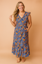 Flowy ankle-length blue and orange floral dress with flutter sleeves, a V-neckline, and a relaxed waistline, styled with strappy flat sandals.