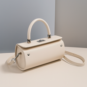 Smooth off-white cylindrical handbag with silver hardware, top handle, and detachable strap on a neutral platform.