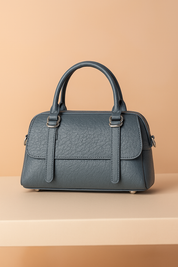 Structured blue-grey pebbled leather handbag with dual top handles, vertical strap detailing, and silver-tone hardware on a pastel sand-to-pink gradient background, styled on a neutral table platform.