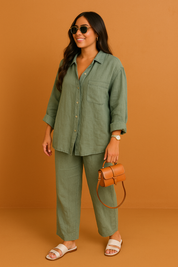 Sage green button-up linen shirt and matching straight-leg pants styled with white sandals and a tan mini handbag, posed against a smooth orange sand-toned studio backdrop.