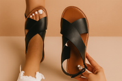 Women's black cross-strap flat sandals with a tan sole and decorative pearl zipper detail at the heel, crafted in a matte leather-like finish with white contrast stitching.
