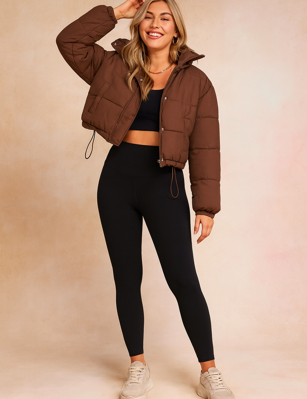 Chocolate brown cropped puffer jacket with front snap buttons and drawstring hem, paired with a black fitted crop top, high-waisted black leggings, and beige chunky sneakers.