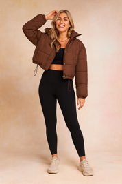 Chocolate brown cropped puffer jacket with front snap buttons and drawstring hem, paired with a black fitted crop top, high-waisted black leggings, and beige chunky sneakers.