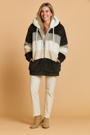 Cozy oversized faux fur hooded coat with color block design in black, grey, white, and beige, paired with cream straight-leg pants and beige ankle boots.