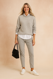 Light grey oversized knit sweater layered over a crisp white collared shirt, paired with slim-fit light grey trousers, white sneakers, and a structured black suede handbag.