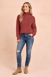 Rose-colored cable-knit turtleneck sweater with intricate central twist pattern, paired with distressed blue jeans and tan suede ankle boots.