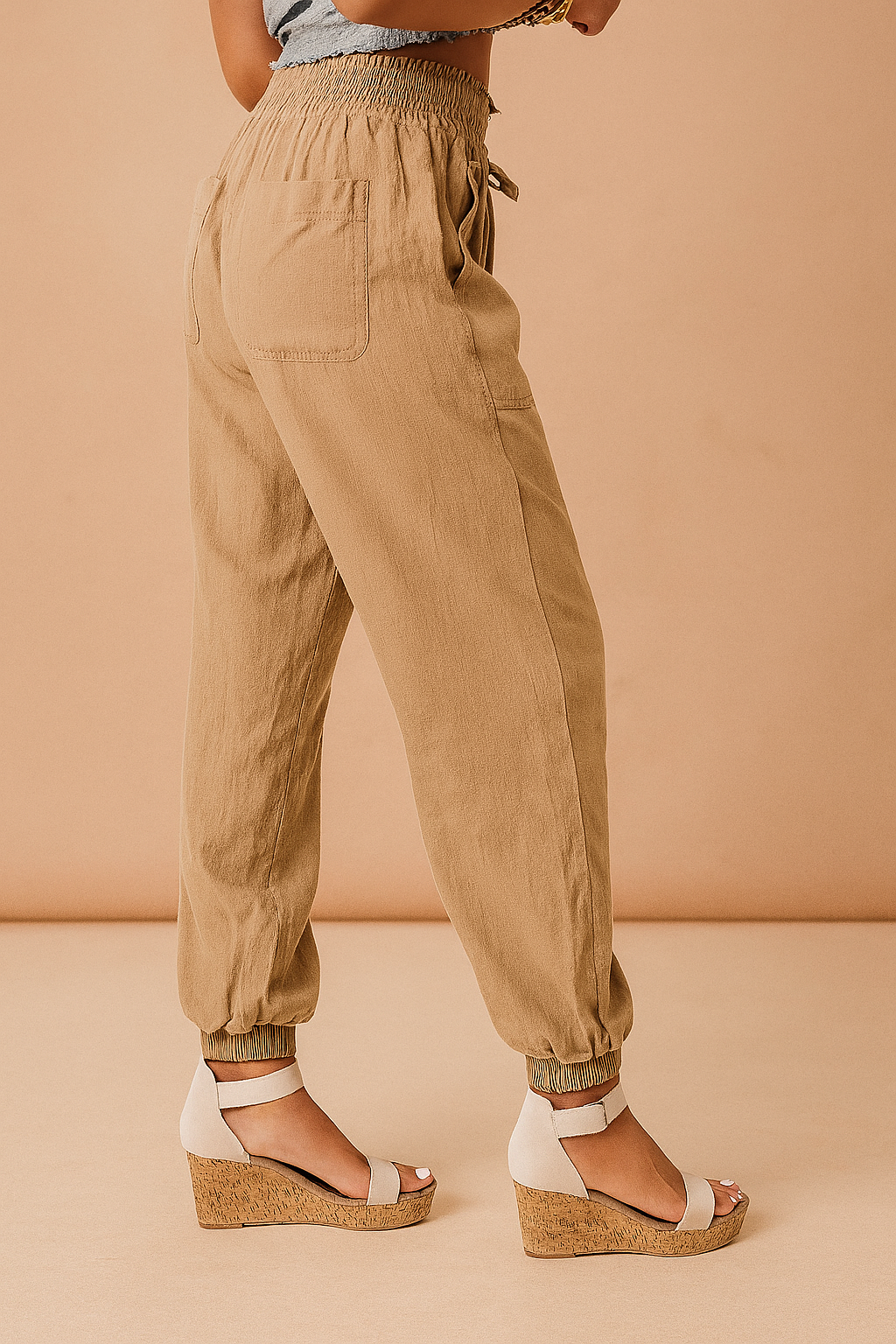 Tan linen jogger pants with elastic waistband, side pockets, and gathered ankle cuffs, styled with white ankle-strap wedge sandals.