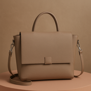 Taupe leather handbag with structured design, front flap closure, and adjustable strap, positioned on a soft salmon-toned platform with a warm beige gradient background.