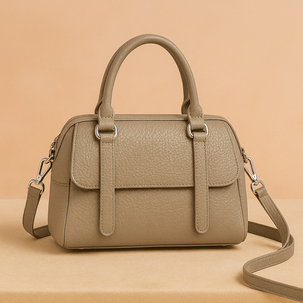 Taupe-colored pebbled leather handbag with rounded handles, front flap pocket, and silver hardware, displayed on a soft beige surface with warm gradient lighting.