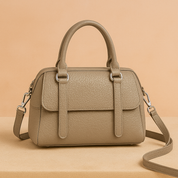 Taupe-colored pebbled leather handbag with rounded handles, front flap pocket, and silver hardware, displayed on a soft beige surface with warm gradient lighting.