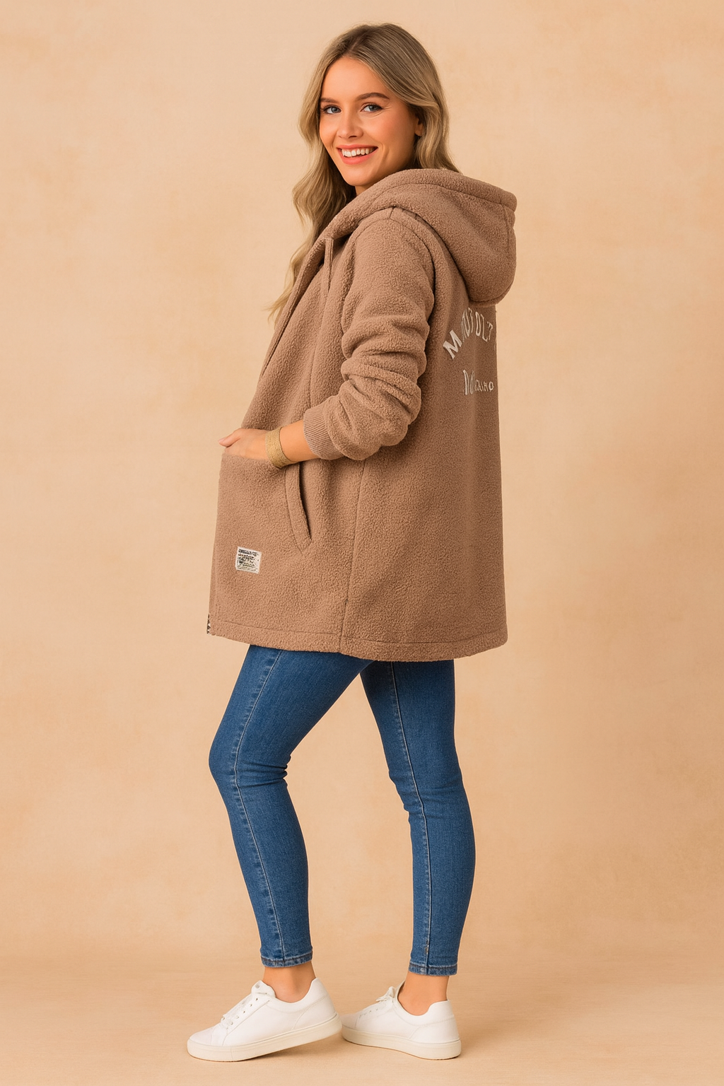 Cozy oversized taupe sherpa hoodie jacket with long sleeves, ribbed cuffs, kangaroo-style pockets, and embroidered back detail. Styled with fitted blue jeans and white sneakers for a casual winter look.
