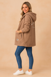 Cozy oversized taupe sherpa hoodie jacket with long sleeves, ribbed cuffs, kangaroo-style pockets, and embroidered back detail. Styled with fitted blue jeans and white sneakers for a casual winter look.