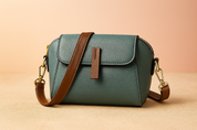 Teal pebbled leather crossbody bag with brown strap, flap closure, and gold hardware, styled on a sand-tone tabletop with gradient backdrop.