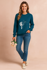 Teal long-sleeve sweatshirt with white dandelion and hot air balloon graphic, paired with fitted blue jeans and beige sneakers.