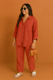 Terracotta red button-up linen shirt paired with matching straight-leg pants, styled with white slide sandals and a structured tan mini handbag, posed in a warm-toned studio.