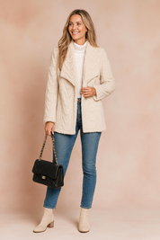 Cream-textured faux fur knit jacket with chevron pattern, worn over a white ribbed turtleneck top and paired with slim-fit mid-wash jeans and beige block-heel ankle boots.