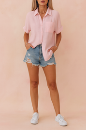 Loose-fit pastel pink button-up shirt with textured vertical pleats, styled with rolled sleeves and a chest pocket, paired with distressed denim shorts and white sneakers—perfect for casual summer looks.