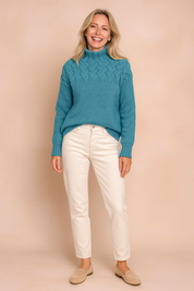 Turquoise cable-knit high-neck sweater with intricate braided pattern on the upper bodice, paired with off-white straight-leg pants and beige suede loafers.