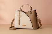 Two-tone structured handbag in ivory and camel with gold hardware, heart-shaped tassel charm, and matching crossbody strap on a warm sand and pastel blush background.