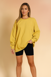 Oversized yellow crewneck sweatshirt with drop shoulders and ribbed cuffs, styled over black ribbed biker shorts, showcased against a softly lit warm sand and pastel pink gradient background in a fashion shoot setting.