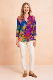 Vibrant abstract floral print long-sleeve blouse in rich shades of magenta, blue, yellow, green, and orange, featuring a classic button-down front and cuffed sleeves, paired with fitted white trousers and tan suede loafers.