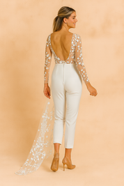 Elegant white bridal jumpsuit with sheer long sleeves, floral lace embroidery, and a flowing tulle train featuring delicate flower appliqué details.
