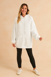 White cable knit hoodie with kangaroo pocket and drawstring, paired with black fitted pants and casual sneakers.