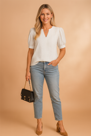 White short-sleeve eyelet blouse with puffed sleeves, vertical embroidery detailing, and modest V-neck, styled with light blue cropped jeans, a black handbag, and tan suede block heels.