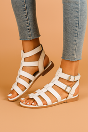 White faux leather gladiator sandals with multiple horizontal straps, dual silver buckles at the ankle, and a low-profile tan sole with visible stitching.