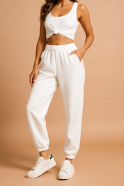 High-waisted white jogger pants with elastic cuffs and side pockets, paired with a knotted white crop top and minimalist white sneakers featuring green heel tabs.