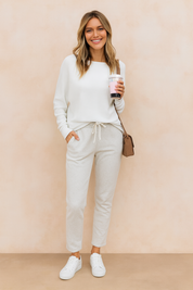 White long-sleeve knit top with a relaxed fit, paired with light grey drawstring sweatpants and classic white sneakers, accessorized with a small brown crossbody bag.
