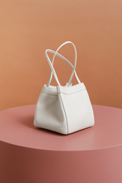 Compact white leather handbag with pebbled texture and double thin handles, placed on a blush pink circular base with a warm peach-to-orange gradient backdrop.