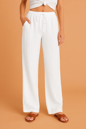 White linen wide-leg drawstring pants featuring a relaxed fit and soft texture, styled with brown sandals for a breezy summer look, set against an orange sandy backdrop