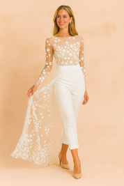 Elegant white bridal jumpsuit with sheer floral lace bodice, long lace sleeves, fitted ankle-length trousers, and a flowing floral lace overlay train, styled with nude pointed-toe heels.
