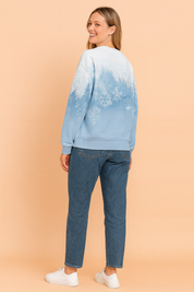 Light blue gradient sweatshirt with white frosted pine branch and snowflake print on the back, paired with mid-wash straight-leg jeans and white lace-up sneakers.
