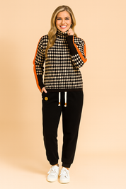 Oversized houndstooth turtleneck sweater with bold orange stripe detailing on sleeves, paired with black jogger pants and white sneakers.