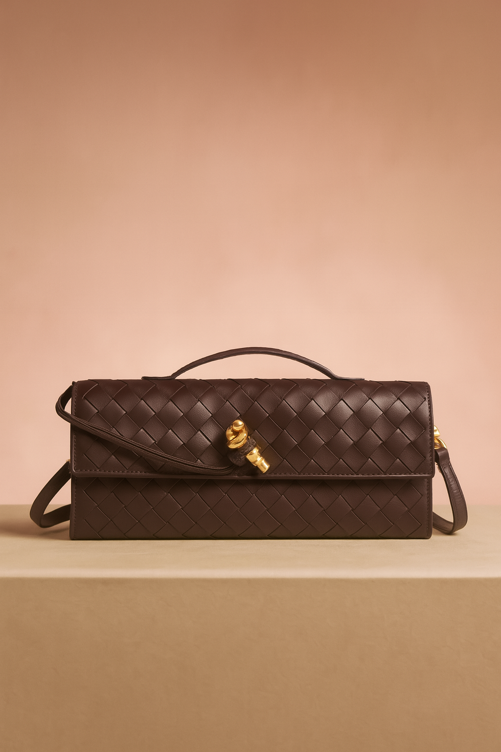 Rectangular dark brown woven leather handbag with a top handle, long shoulder strap, and gold hardware clasp, displayed on a neutral platform with soft gradient background.