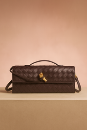 Rectangular dark brown woven leather handbag with a top handle, long shoulder strap, and gold hardware clasp, displayed on a neutral platform with soft gradient background.