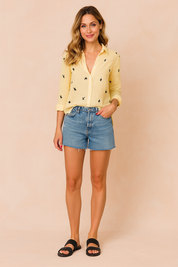 Long-sleeve yellow button-up blouse with black bird print, styled with high-rise frayed denim shorts and black sandals.